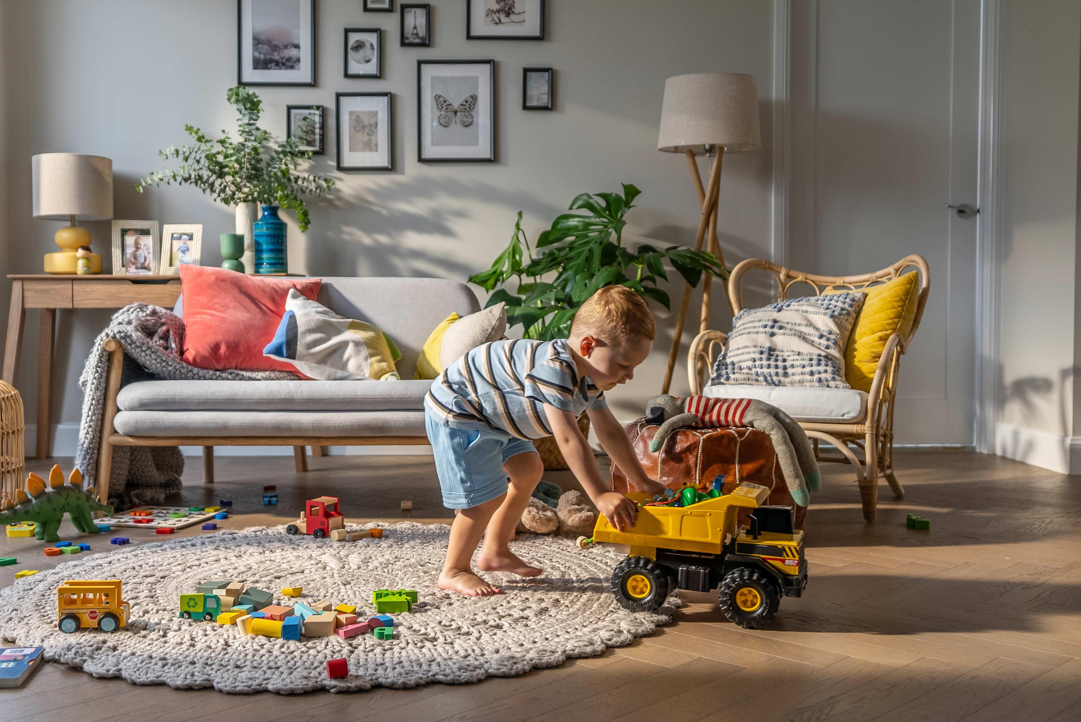 Young child playing with a toy dump truck in a cozy, well-decorated living room with toys scattered on a textured rug and a gallery wall in the background.