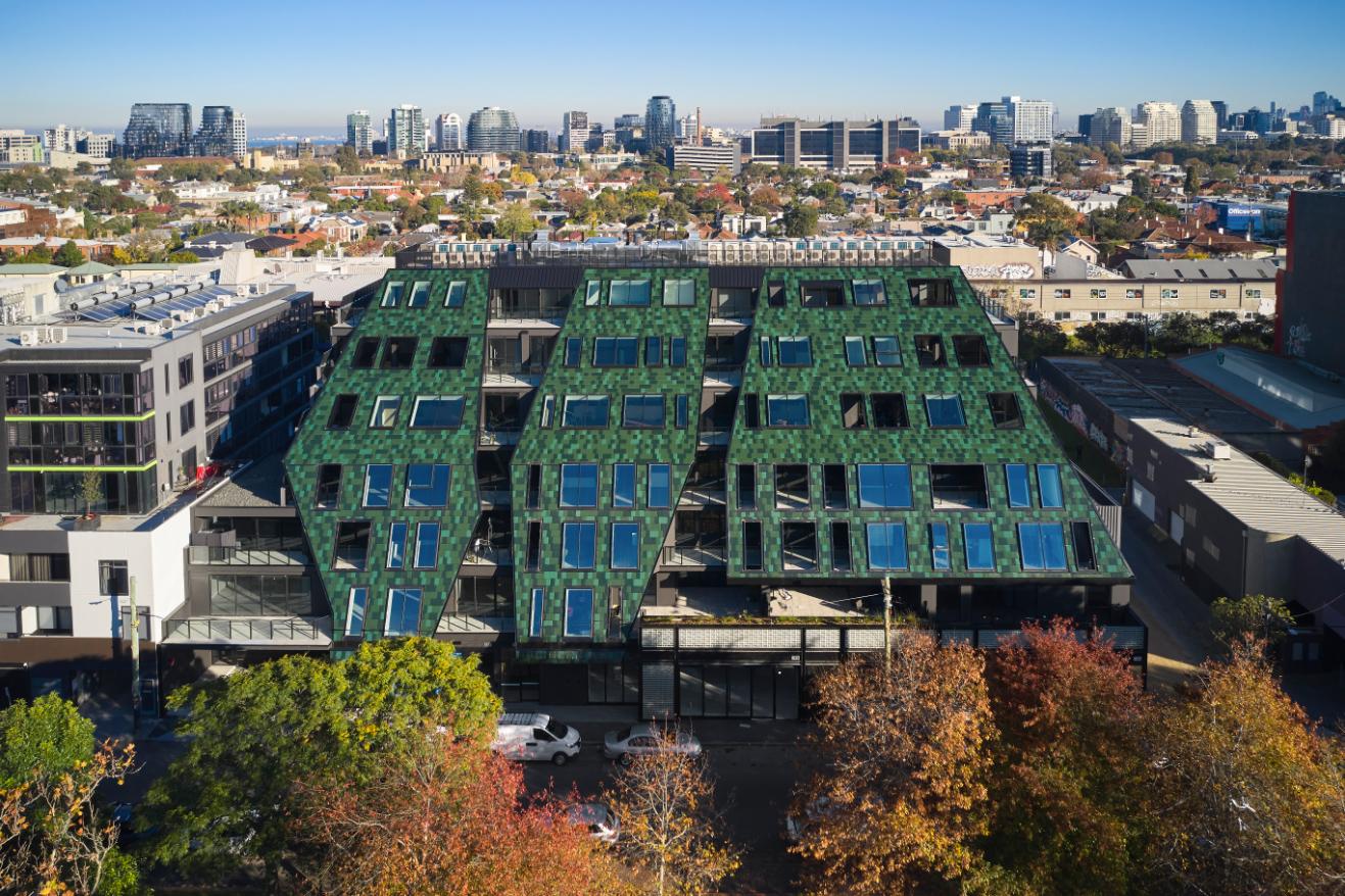 Aerial view of a modern green-roofed building surrounded by urban landscape and trees, with city skyline in the background.