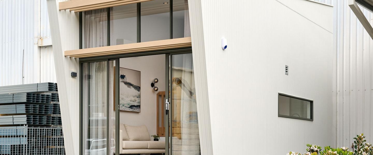 Modern building exterior with large glass sliding doors, revealing a cozy interior with a couch and wall art.