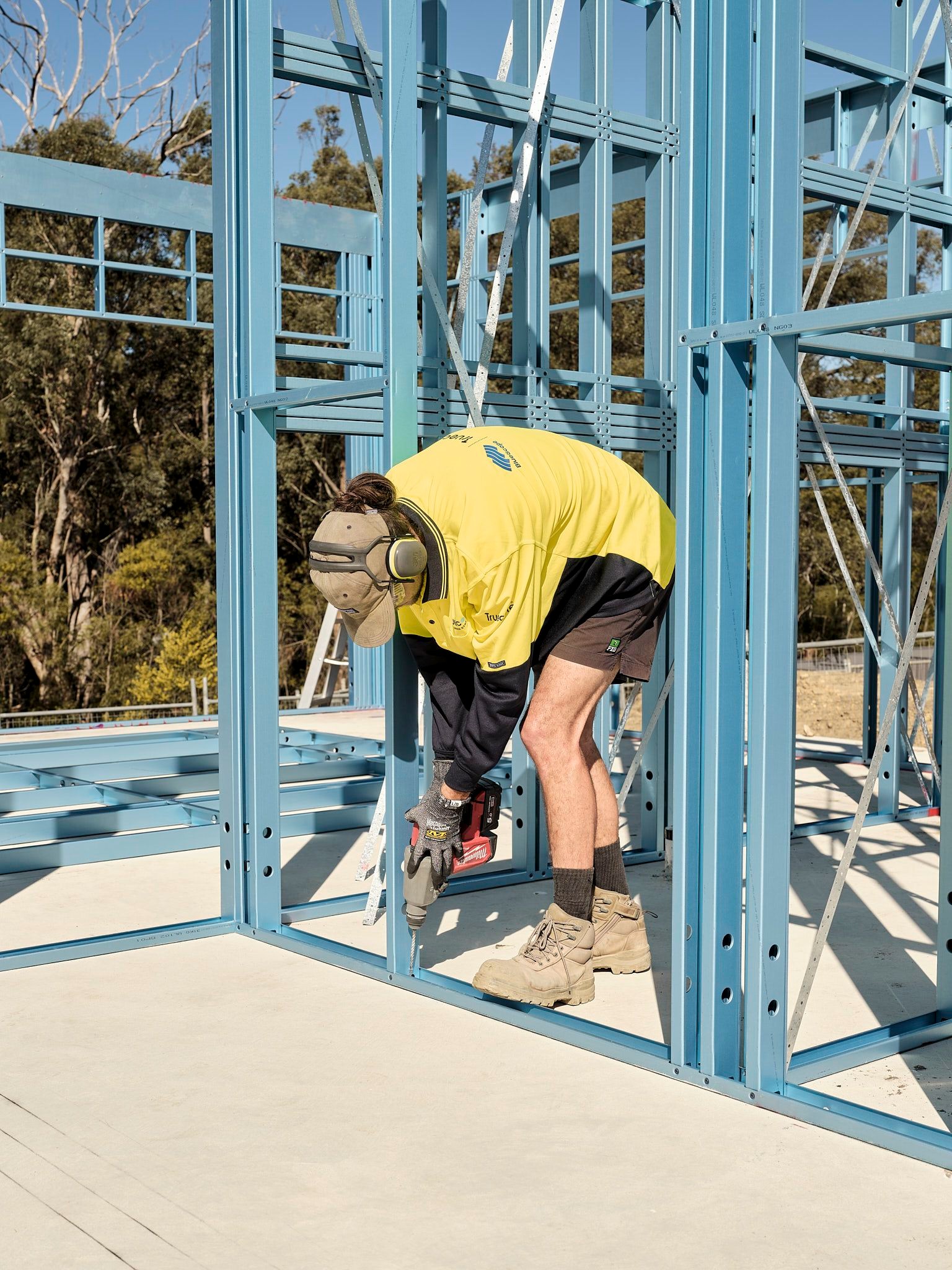 Construction worker in protective gear assembling a steel frame structure on a building site, using a power drill for securing connections.
