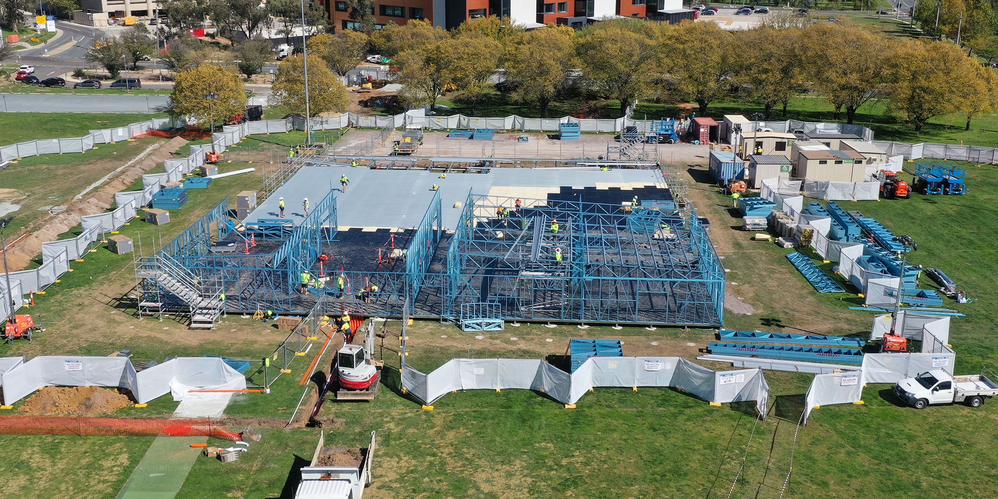 A construction site featuring the early stages of a building framework made from blue steel. Workers in high-visibility clothing are scattered around the site, and scaffolding surrounds the steel frame structure. The site is enclosed with temporary fencing, and several construction vehicles and equipment are visible. The surrounding area includes trees and an adjacent building in the background.