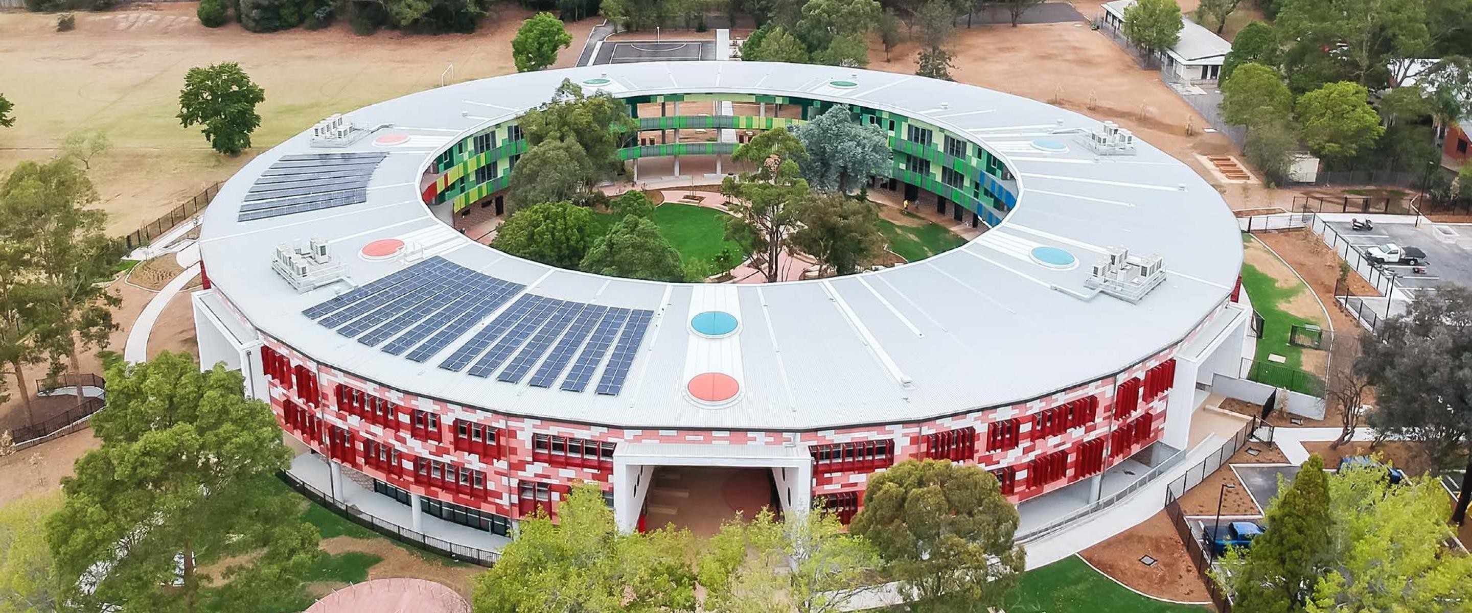 Aerial view of a circular modern building with a large open courtyard at its center. The building features colorful green and blue panels on one side and red panels on another, with a rooftop equipped with solar panels. The courtyard includes landscaped greenery and trees, creating a vibrant and sustainable architectural design. Surrounding the building are pathways, parking areas, and open fields.