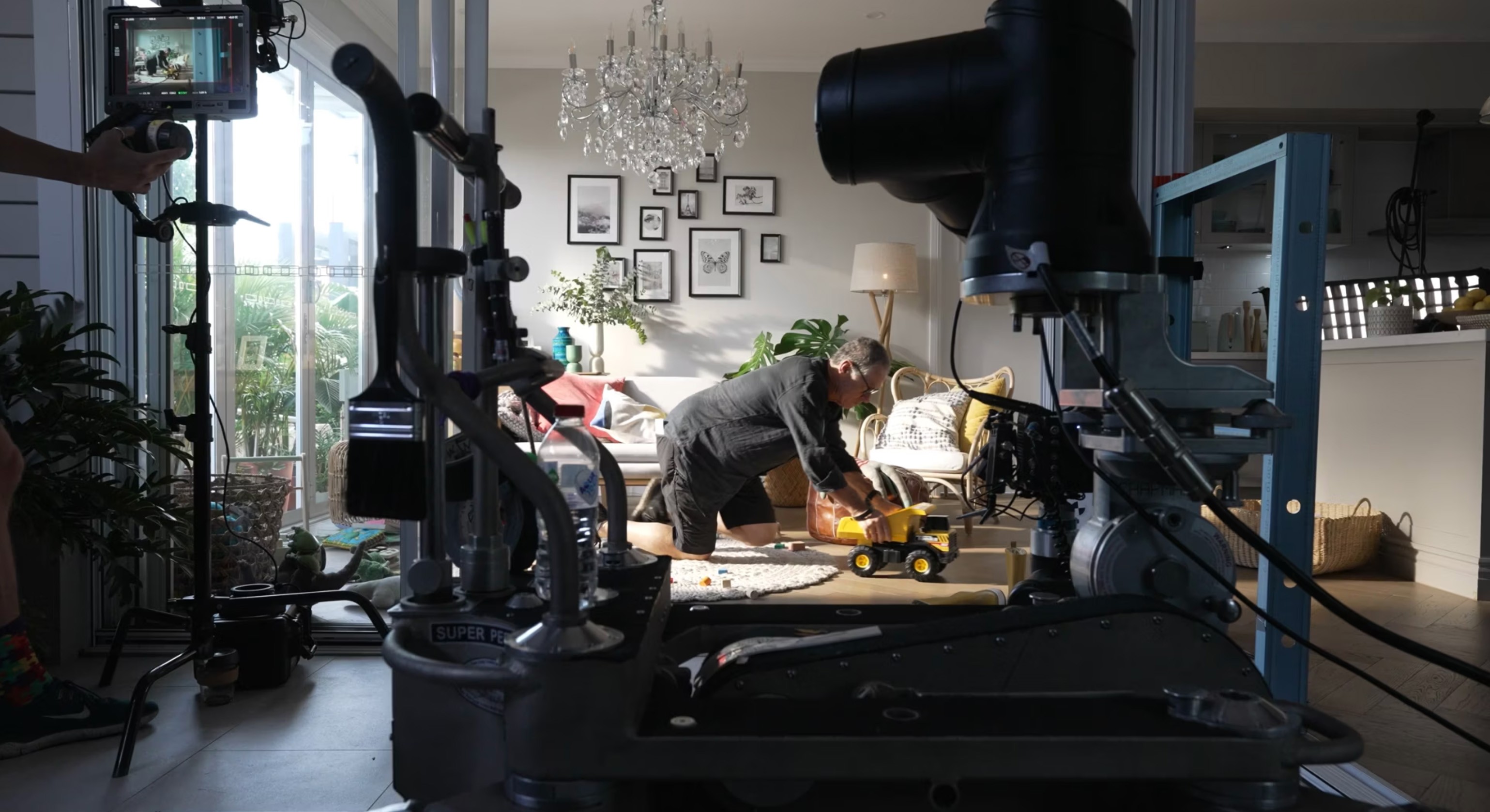 Behind-the-scenes shot of a video production set inside a well-lit modern living room. A man is kneeling on the floor, playing with a yellow toy dump truck, while a professional camera rig and lighting equipment capture the scene. A monitor displays the live feed of the shoot. The setting includes a chandelier, framed wall art, indoor plants, and cozy furniture, creating a warm and inviting atmosphere.