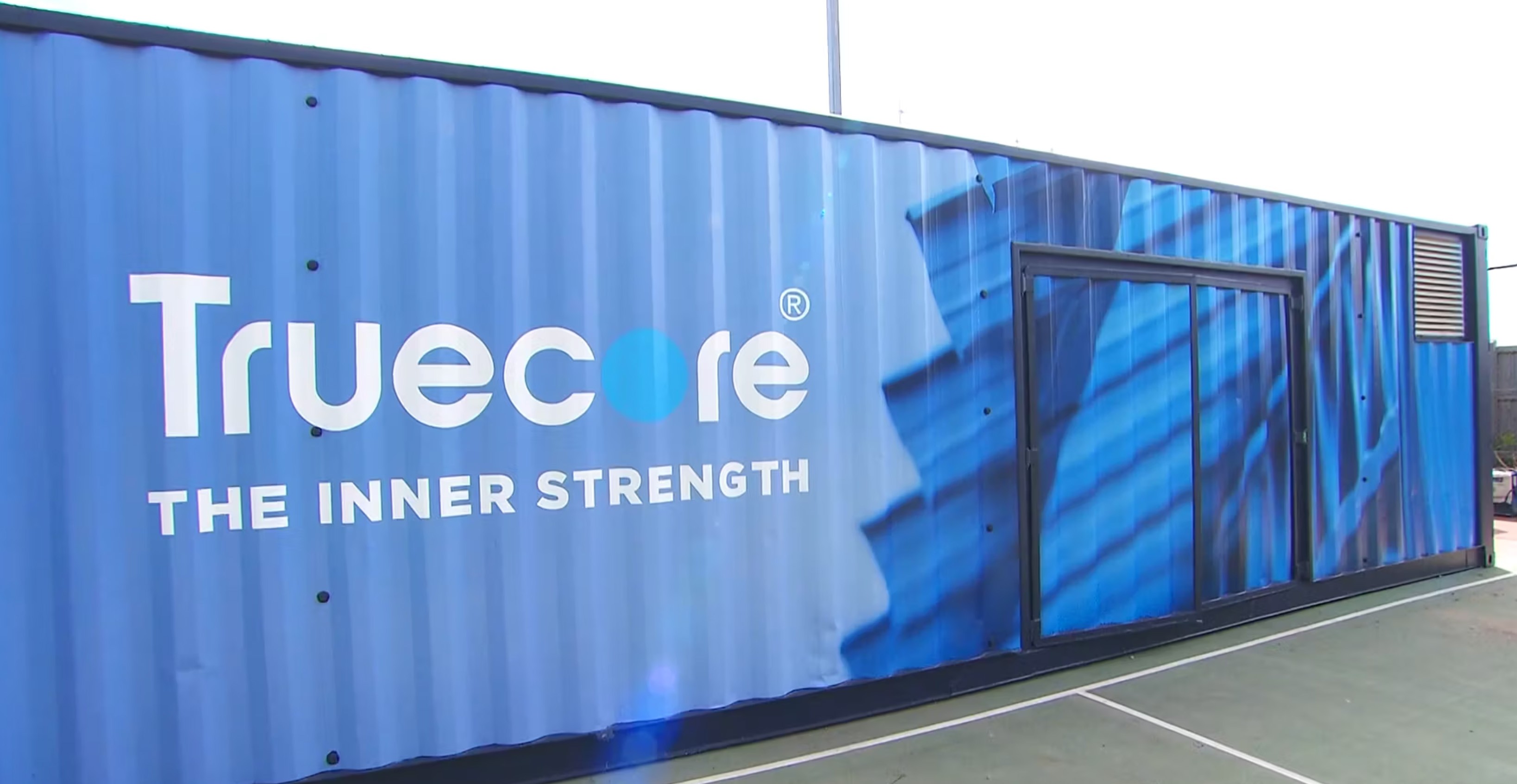 A shipping container repurposed as a promotional display for Truecore® steel, featuring a bold blue exterior wrap with the Truecore logo and the slogan " The Inner Strength". The design includes an abstract image of steel framing, emphasizing the product's structural durability. The container is placed on a sports court with a sliding door entrance and a ventilation panel on one side.